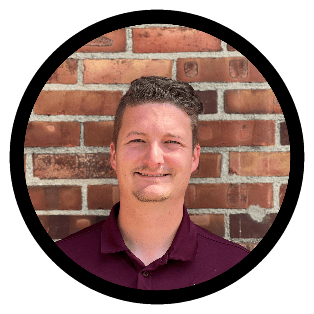A headshot of a brunette man wearing a maroon polo shirt while standing against a brick wall.